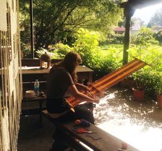 a woman sitting on a bench working on an item in her yard with sunlight streaming through the trees