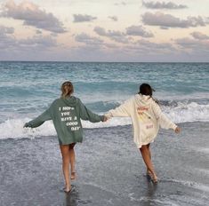 two young women walking on the beach holding hands and looking out at the ocean while wearing sweatshirts