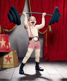 a young boy is performing with a barbell in front of red curtained curtains