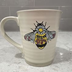 a ceramic mug with a bee painted on the side and flowers in the middle, sitting on a marble countertop