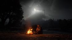 a man sitting in front of a campfire under a dark sky with the moon behind him