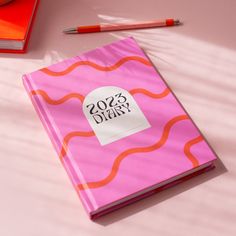 a pink notebook sitting on top of a table next to a red pen and pencil