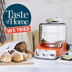 an orange and white food processor sitting on top of a counter next to bread rolls