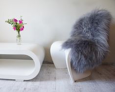 a white table with a vase and flowers on it next to a chair that has a gray fur cushion