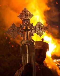 a man holding up a cross in front of a fire that is burning behind him