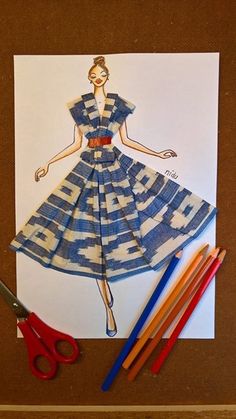 a drawing of a woman in a blue dress on paper next to scissors and colored pencils