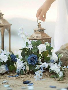 a close up of a person placing flowers in a vase on the sand near two lanterns