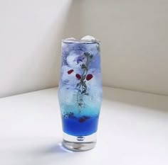 a glass filled with blue liquid and flowers on top of a white table next to a wall