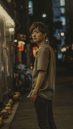 a man standing on the sidewalk in front of a building at night with his head turned to the side