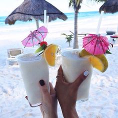 two people holding up drinks on the beach with umbrellas in the sand behind them