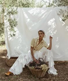 a woman sitting on the ground next to a basket full of grapes in front of a white curtain