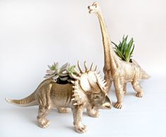 two toy dinosaurs with plants in their mouths on a white background, one is gold and the other is silver