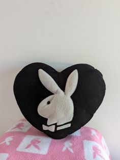 a black heart shaped pillow with a white rabbit on it's face sitting on top of a pink and white blanket