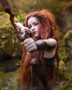 a woman with long red hair holding a bow and arrow in her hands while standing on mossy ground