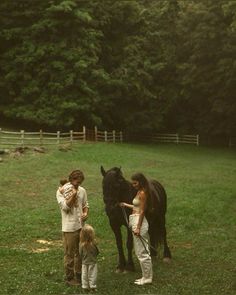 two women and a child are standing next to a black horse in a grassy field