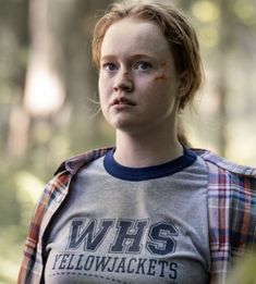 a woman with red hair wearing a grey shirt and plaid vest in the woods looking at the camera