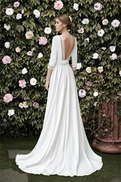 a woman standing in front of a flower covered wall wearing a white dress with open back