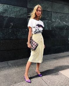 a woman in a skirt and t - shirt is standing on the sidewalk with her handbag