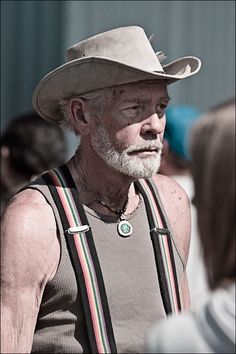 an old man wearing a hat and suspenders is standing in front of other people