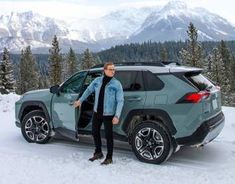 a man standing next to a car in the snow with mountains in the back ground