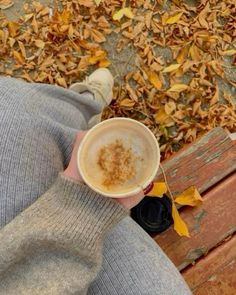 a person sitting on a bench holding a cup of coffee in their hand with leaves around them