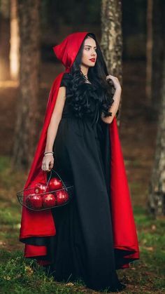 a woman in a red cape and black dress holding an apple basket standing in the woods