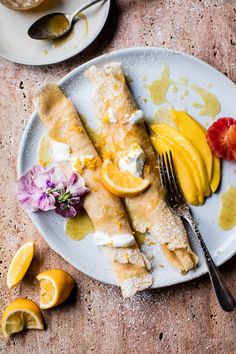 a white plate topped with crepes covered in lemon and whipped cream next to sliced fruit