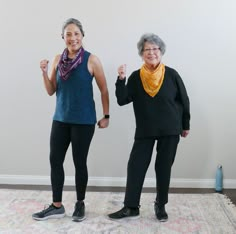 two older women standing next to each other in front of a white wall and rug