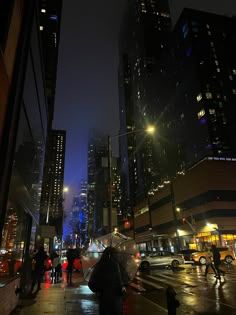 people are walking down the street at night with umbrellas in hand and buildings lit up behind them