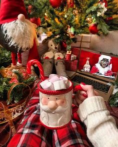 a person sitting in front of a christmas tree holding a coffee mug with santa clause on it