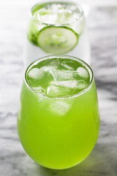 two glasses filled with green liquid and cucumber slices on the rim, sitting on a marble surface