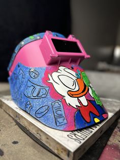 a pink and blue helmet sitting on top of a piece of wood with graffiti painted on it