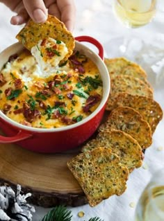 a hand dipping a cracker into a red casserole dish with bacon and cheese