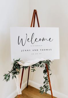 a welcome sign on top of a easel in front of a white wall with greenery