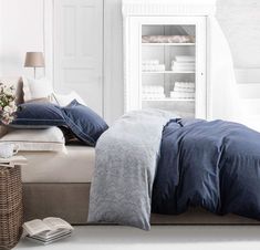 a bed with blue sheets and pillows in a white room next to a book shelf