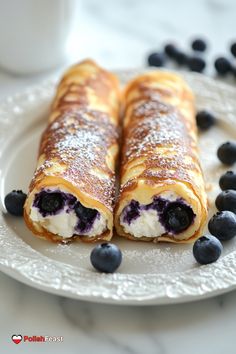 two blueberry crepes on a white plate with powdered sugar and blueberries