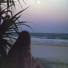 a woman is sitting on the beach watching the ocean at dusk with her back turned to the camera