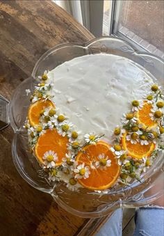 a cake decorated with oranges and daisies on a glass plate in front of a window