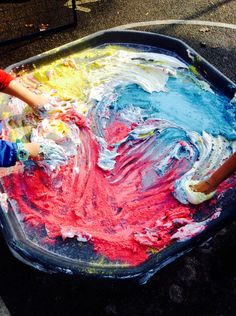 two children are painting with colorful paints on a large metal tray in an outdoor area