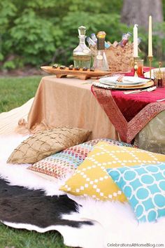 a table set up with pillows, plates and candles on top of a blanket in the grass