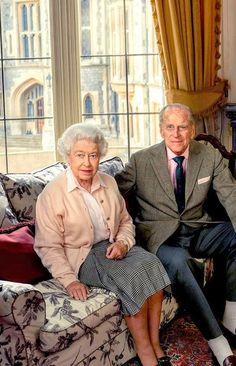 an older man and woman sitting on a couch in front of a window with curtains