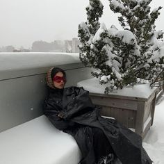 a woman sitting on top of a bench covered in snow next to a potted tree