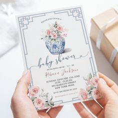 a person holding up a baby shower card with flowers on it and a gift box in the background