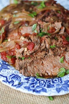 a blue and white plate topped with meat, onions and tomato sauce on top of a woven table cloth