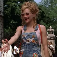 a woman in overalls and sun glasses walking down the street with her hand out