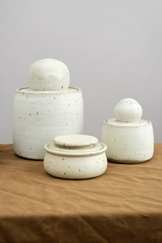 three white ceramic containers sitting on top of a brown tablecloth covered table next to each other