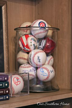 some baseballs are in a glass container on a shelf next to books and a clock