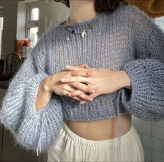 a woman with her hands on her chest wearing a blue knitted sweater and white pants