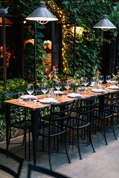 an outdoor dining table set up with wine glasses and place settings in front of greenery