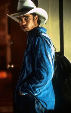 a man with a cowboy hat standing in front of a wall and looking at the camera